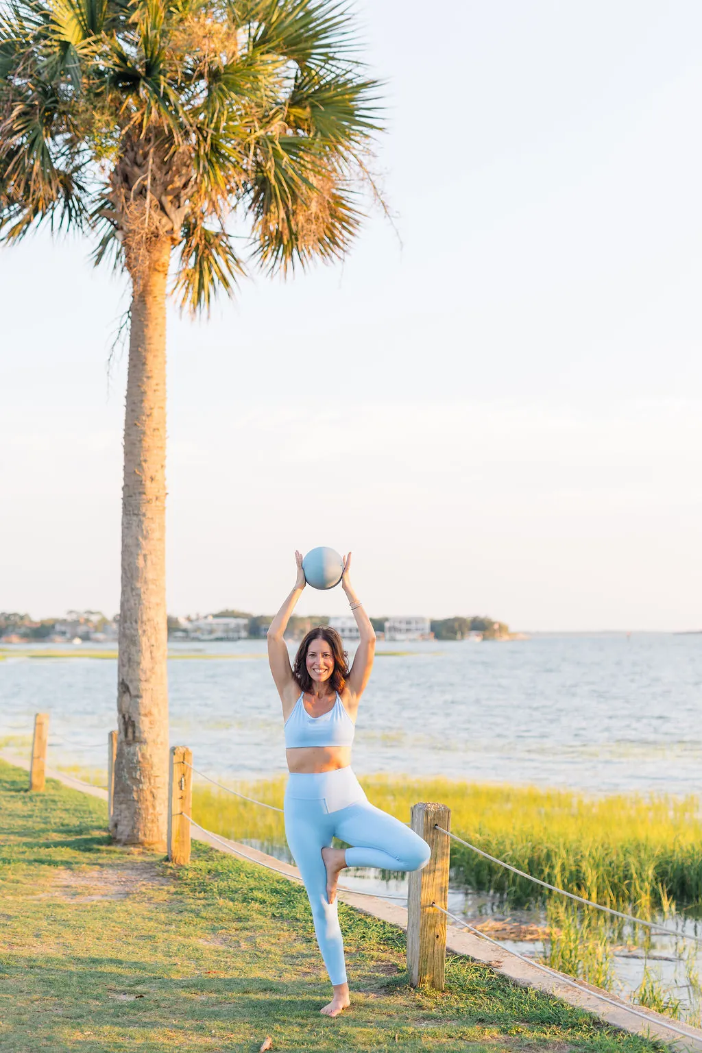 Instructor balancing in tree pose with a pilates ball overhead beneath a palmetto tree by the harbor